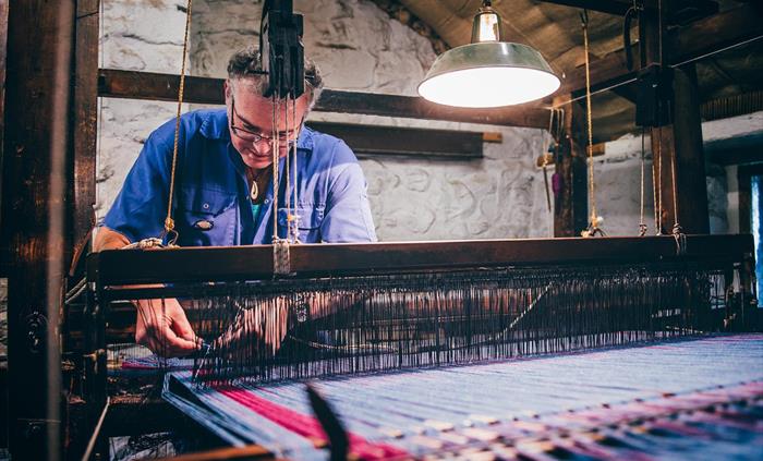 a man in blue overalls works at a traditional loom weaving a blanket